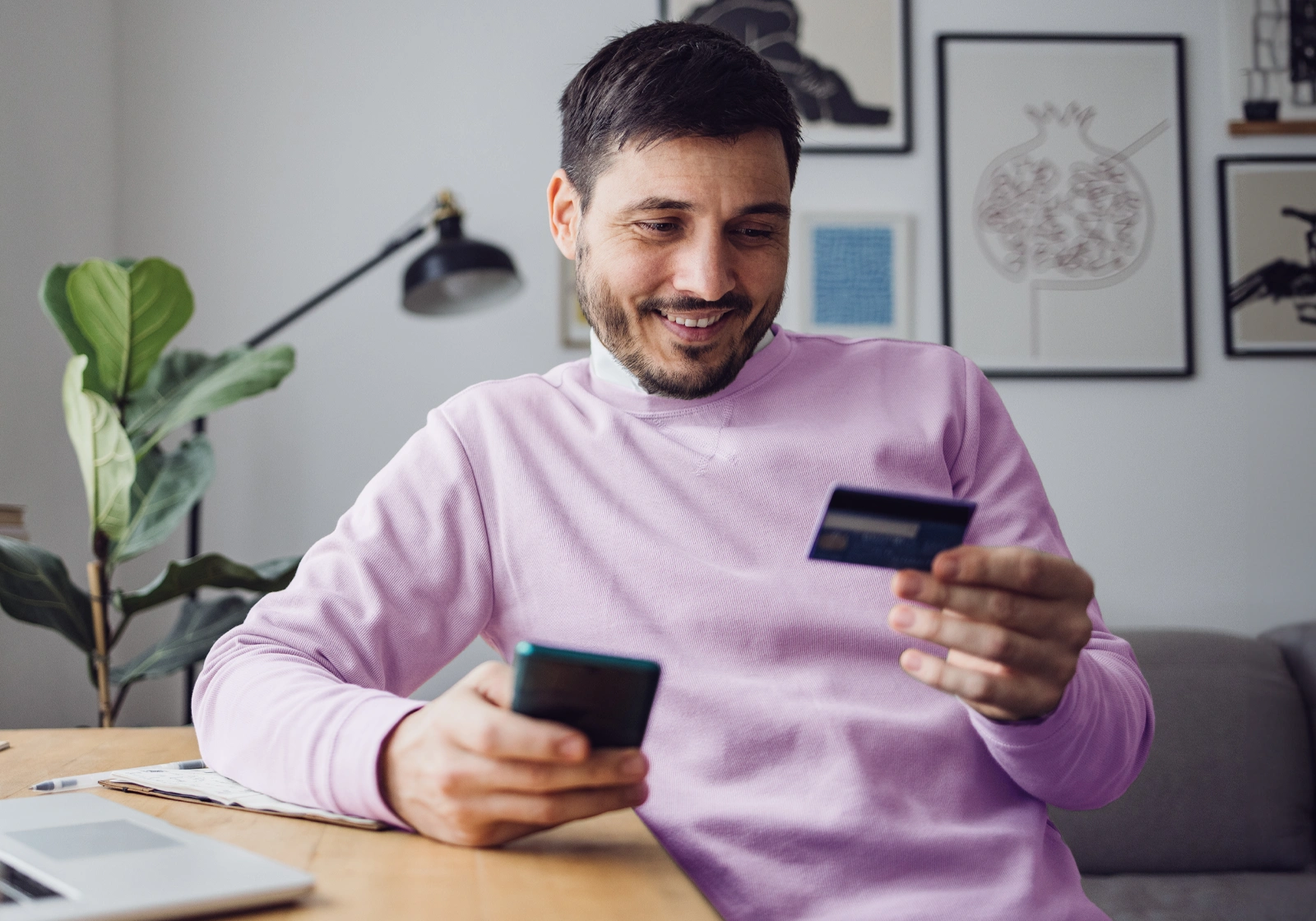 A man sits at a desk in his home with a credit card in one hand while using a smartphone to manage his account.