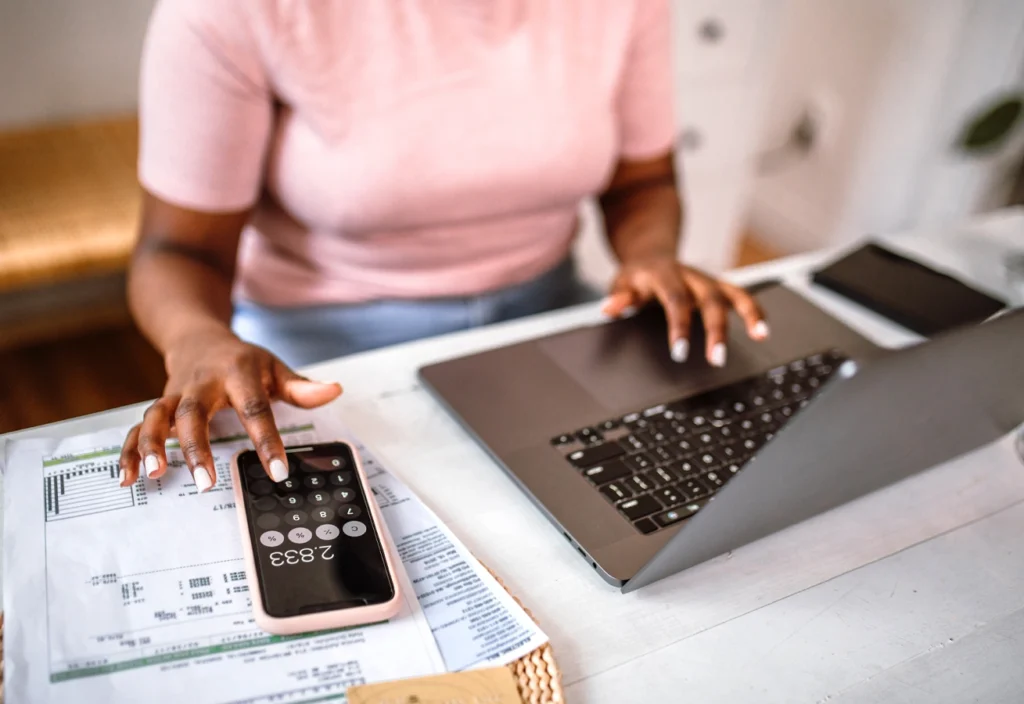 A person uses a smartphone calculator as they perform budgeting tasks on a laptop.