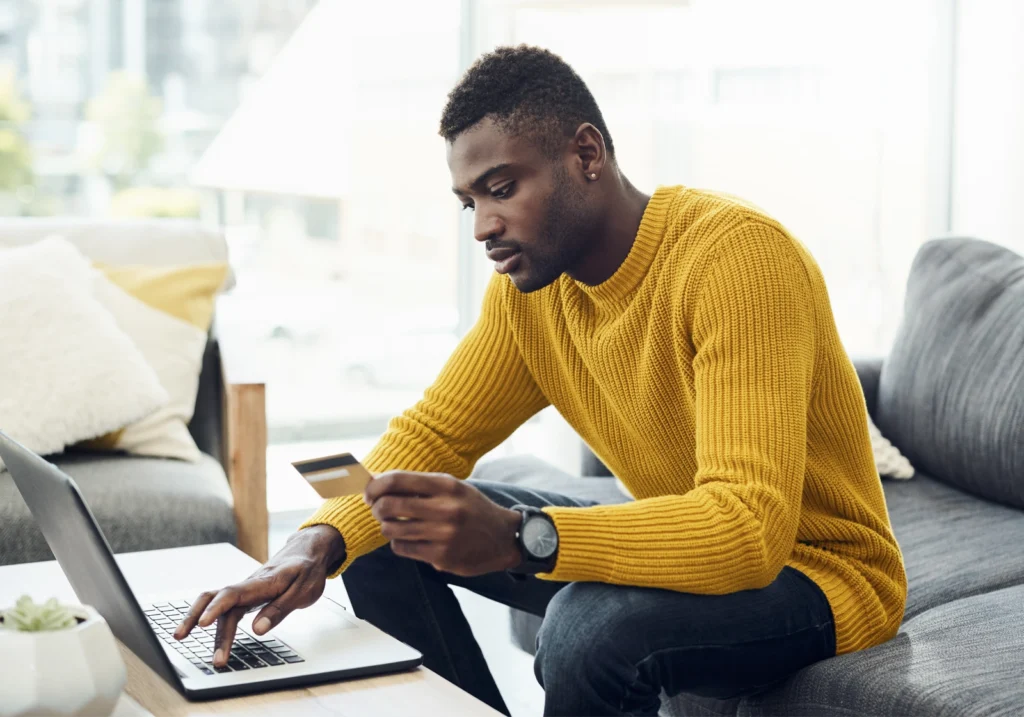 A man sits on his couch and holds a credit card while using a laptop to manage his account.