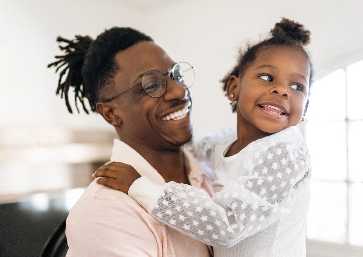 A confident adult smiles while holding a child at home.