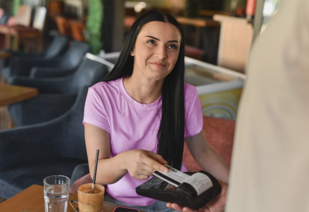 A woman taps her credit card against a card reader to pay for a purchase in a cafe.