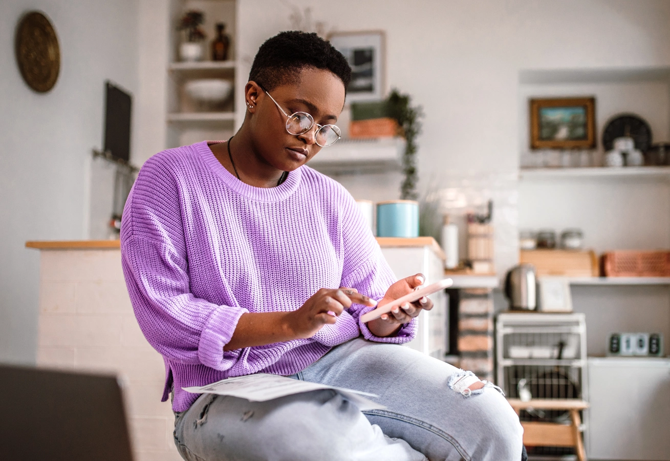 A person wearing glasses sits in their home and uses a smartphone to research money management tips.