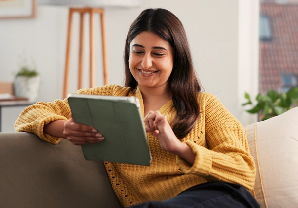 A woman sits on her couch and uses a tablet to review her debt progress.