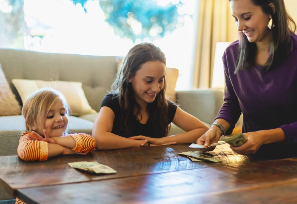 A smiling mother hands out allowance to her two daughters after talking about money with them.